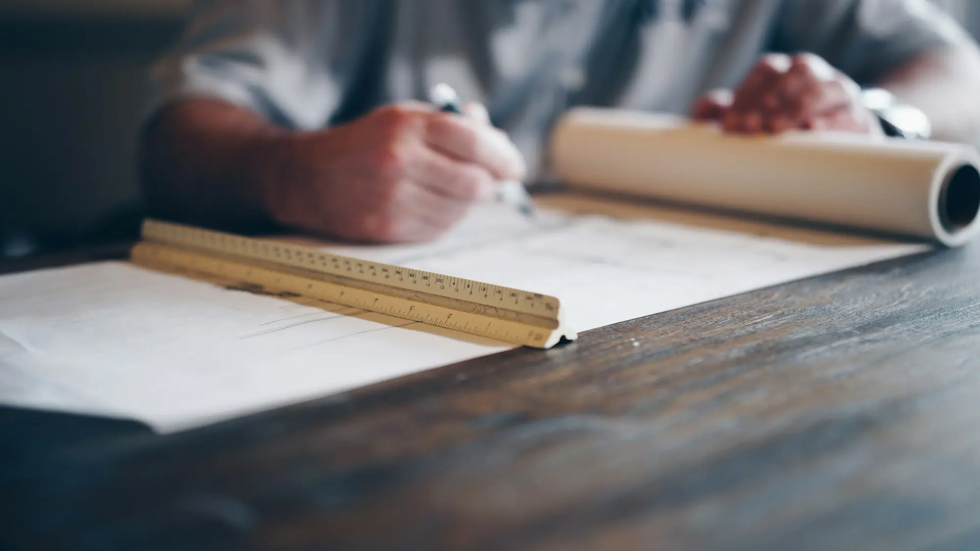 Architect sketching building plans at a timber desk with a scale ruler and a roll of drawings