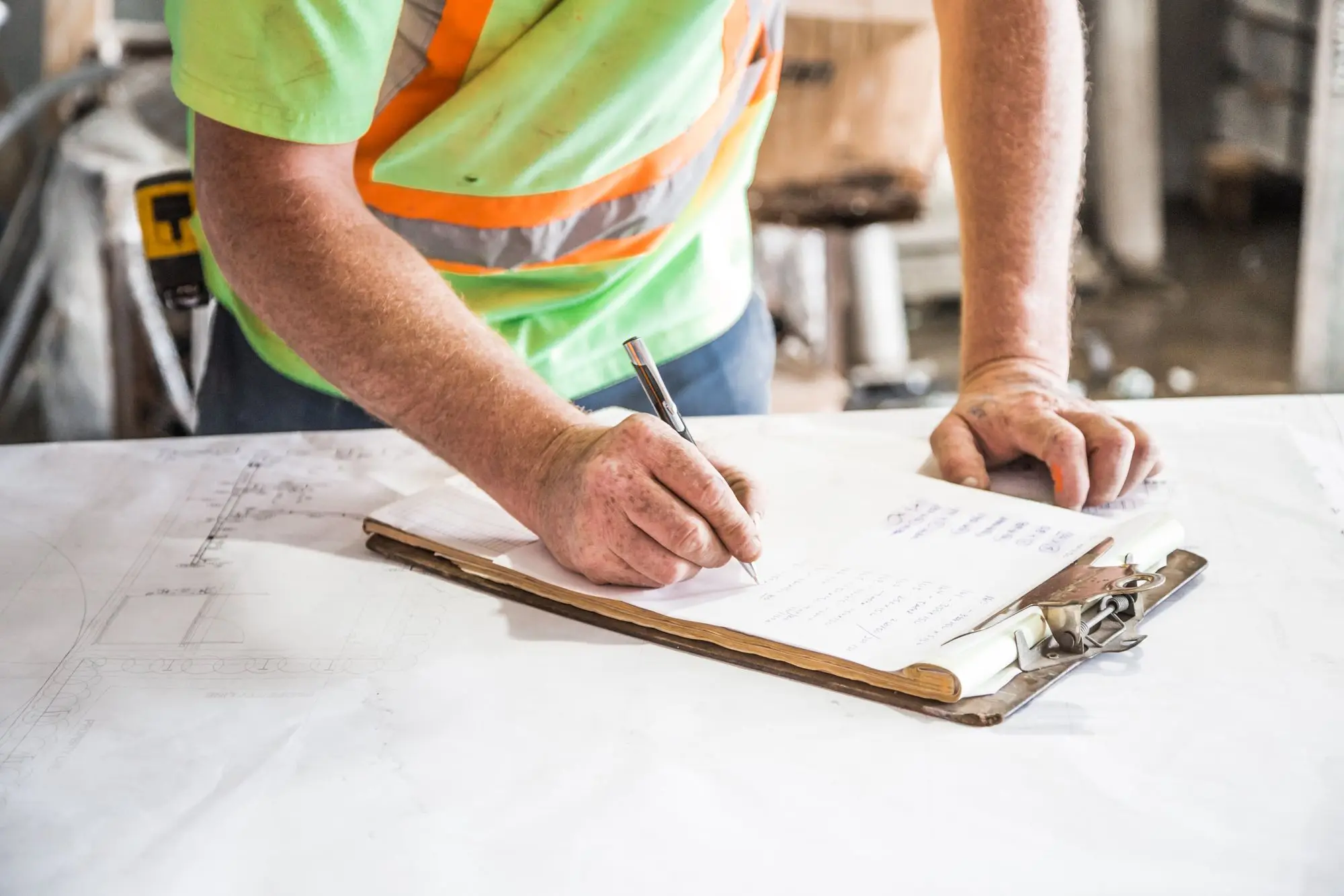 Builder in a hi-vis work shirt writing figures on a clipboard laid over a set of building plans